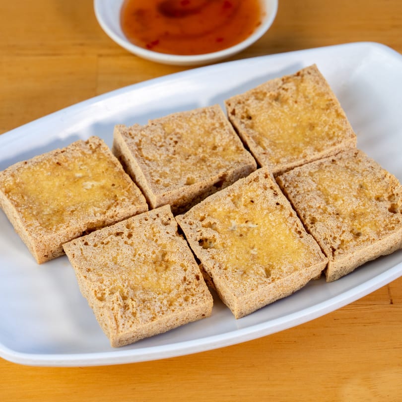 Six pieces of crispy fried tofu on a white rectangular plate with a small bowl of chili sauce.