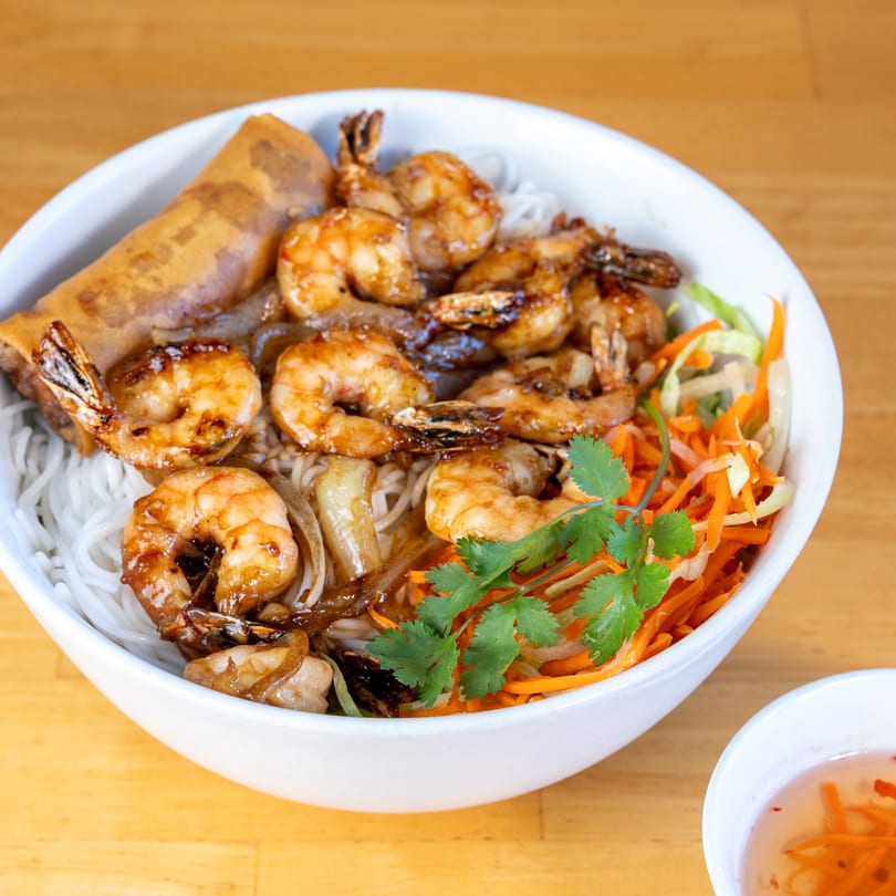 A white bowl of Vietnamese vermicelli noodles with grilled shrimp, egg roll, and fresh vegetables.