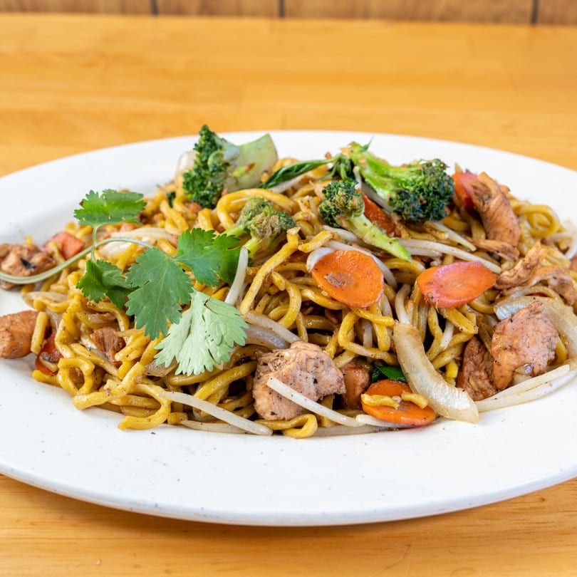 Vietnamese stir-fried noodles with chicken, broccoli, carrots, onions, and cilantro on a white plate.